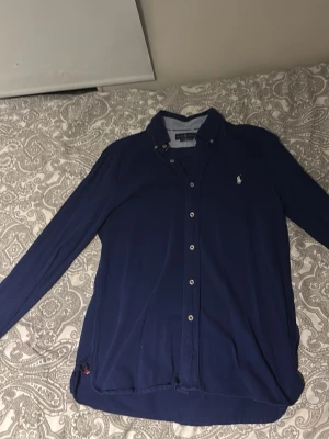 Blå skjorta från Ralph Lauren M - Säljer en snygg blå skjorta från Ralph Lauren i modellen Featherweight Mesh. Skjortan har klassisk krage, knappar hela vägen och lång ärm. Ikoniska loggan broderad på bröstet. Perfekt för dig som gillar stilrena plagg med premiumkänsla.