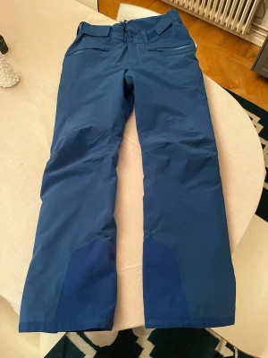 Skidbyxor från Bergans of Norway - Säljer ett par blå skidbyxor från Bergans of Norway, modell Oppdal. Byxorna har dragkedjor på sidorna, förstärkta partier vid bensluten och breda hällor för bälte. Tillverkade i slitstark polyester med isolerande foder, perfekta för kalla dagar i skidbacken.