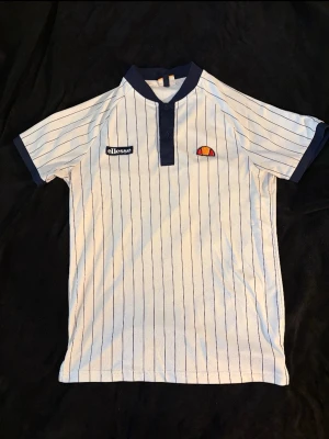 Vit randig t-shirt från Ellesse - Snygg vit t-shirt från Ellesse med tunna marinblå ränder, mörkblå krage och ärmslut. Klassisk knappslå framtill och broderad logga på bröstet. Perfekt för dig som gillar sportig retrostil.