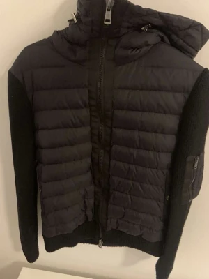 Svart Moncler pufferjacka med stickade ärmar - Säljer en svart hybrid jacka från Moncler med stickade ärmar och huva. Nypris 12000