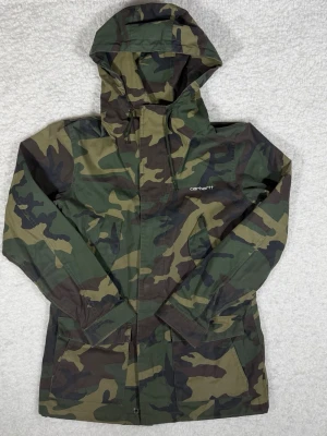 Camouflage parka från Carhartt - Säljer en grön och brun camouflage vindjacka från Carhartt med huva och stora fickor framtill. Jackan har dragsko i huvan, knappar på fickorna och klassisk Carhartt-logga på bröstet. Perfekt för dig som gillar streetwear och utility vibes.