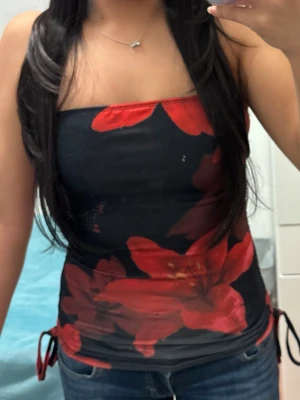 Svart och röd blommig tubtopp - Säljer en snygg tubtopp i svart med stora röda blommor. Toppen är tight och har snörning i sidorna för en extra detalj. Perfekt till jeans eller kjol för en cool look. Materialet känns stretchigt och följsamt.