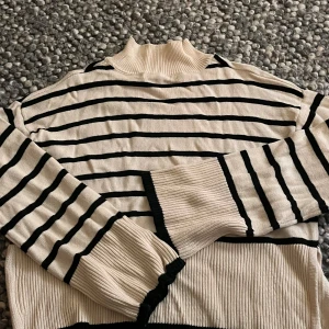 Cream & Black Striped Knit Sweater – Mock Neck – Cozy Minimal Style - Säljer en beige och svart randig stickad tröja från Gina Tricot i storlek S. Tröjan har hög krage, långa ärmar och ribbade muddar samt slits i sidan. Perfekt för dig som gillar klassisk stil med en twist.