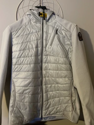 Vit cardigan från Parajumpers - Säljer en snygg vit vindjacka  från Parajumpers med huva och dragkedja framtill. Jackan har en bröstficka med dragkedja, ribbade detaljer på ärmarna och klassisk PJS-patch på axeln. Perfekt för dig som vill ha en stilren och funktionell jacka.