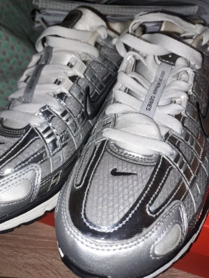 Nike P-6000 silver sneakers - Säljer ett par Nike P-6000 sneakers i silver och vitt med svarta detaljer. Skorna har en futuristisk vibe med mesh och syntetmaterial, samt tydliga svarta swoosh-loggor på sidan och tån. De har vit snörning och en chunky vit och svart sula.
