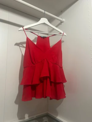 Röd volangig linnetopp - Säljer en röd linnetopp med tunna axelband och dubbla volanger. Toppen har v-ringning och är tillverkad i ett lätt, flödande tyg som ger en luftig känsla. Perfekt för dig som vill ha en färgstark och lekfull look. Som syns på sista bilden har jag klippt bort ett lager.