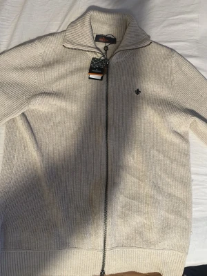 Beige stickad kofta från Morris - Säljer en beige stickad kofta från Morris med hel dragkedja och hög krage. Tröjan har ribbade muddar, diskret Morris-logga broderad på bröstet och snygg detalj på dragkedjan. Perfekt för lager på lager under höst och vinter.