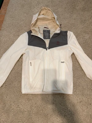 Vit vindjacka från Brunello Cucinelli  - Säljer en lätt vindjacka med huva från Brunello Cucinelli i beige/off-white med grå panel och broderad logo. Tekniskt nylon-material, vattentåligt. I väldigt bra skick, sparsamt använd. Nypris cirka 3200 euro. Inga kvitton kvar. Välkomnar seriösa prisförslag runt