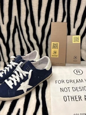 Golden Goose blå sneakers med vit stjärna - Säljer ett par snygga Golden Goose sneakers i marinblå mocka med vit stjärndetalj på sidan och vit häl. Skorna har rund tå, platt sula och vita snören. Märket syns tydligt på hälen och tungan. Perfekta för dig som gillar streetstyle och vill sticka ut. Guldig glittrig där bak som inte synns superstark model av golden goose hör av er vid frågor 