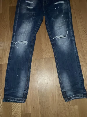 Supply & Demand blå jeans med slitningar - Blå jeans från Supply & Demand med coola slitningar och detaljerade hål vid knäna. Jeansen har en faded effekt och vita färgstänk som ger en edgy vibe. Klassisk femficksmodell och normal passform. Perfekt för dig som gillar streetstyle.