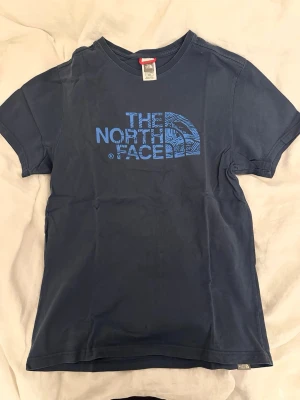Blå t-shirt från The North Face - Snygg mörkblå t-shirt från The North Face med stor logga i blått framtill. Klassisk passform och rund halsringning. Tillverkad i mjuk bomull som känns skön mot huden. Litet hål i nederkant.