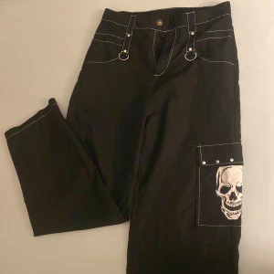 Svarta cargopants med dödskalletryck - Unika svarta cargopants med vit kontrastsöm och stora fickor. Ena benet har ett fett dödskalletryck och nitar på ficklocket. Byxorna har D-ringar i midjan för extra edge och är tillverkade i ett mjukt bomullsliknande material. Perfekta för dig som gillar alternativ stil. Tunnt luftigt material som är väldigt skönt för sommaren.