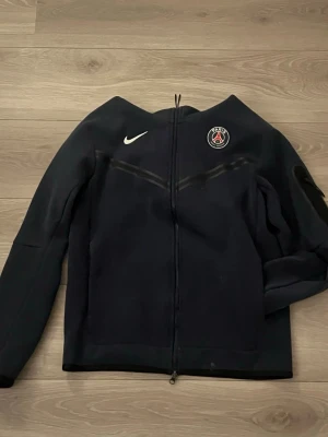 Mörkblått Nike tech set (PSG) - Säljer detta snygga Nike tech set som är broderat med PSG logga. Både byxorna och tröjan är väll använda men inga större defekter, bara lite nopprigt på vissa ställen. Funkar till träning och till vardags. Bara att höra av er vid frågor.