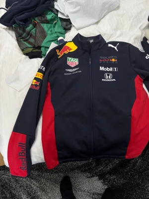 Red Bull Racing jacka från Puma - Säljer en snygg Red Bull Racing jacka från Puma med Aston Martin, Honda och Mobil 1-loggor. Jackan är mörkblå med röda detaljer på sidor och ärmar, dragkedja framtill och flera broderade märken. Perfekt för dig som gillar motorsport och vill sticka ut.