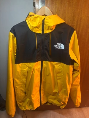Gul och svart vindjacka The North Face - Säljer en snygg gul och svart vindjacka från The North Face i storlek S. Jackan har huva med dragsko, hel dragkedja framtill och logga på bröstet. Tillverkad i lätt polyester, perfekt för blåsiga dagar. Färgerna är riktigt skarpa och jackan har en sportig vibe. Finns nån fläck vid dragkedjan som man ser på sista bilden, kan gå att tvätta bort men inget jag testat. Endast använd några gånger😃