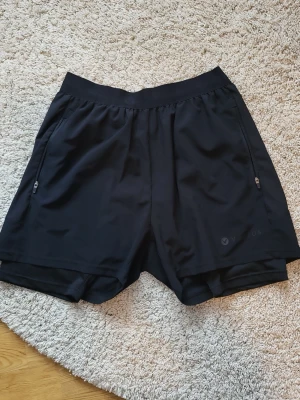 Svarta träningsshorts från Virtus - Säljer ett par svarta träningsshorts från Virtus med elastisk midja och dragkedjeförsedda fickor på sidorna. Shortsen har en innerbyxa för extra komfort och är tillverkade i ett lätt och snabbtorkande syntetmaterial. Perfekta för gymmet eller löprundan.