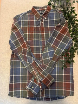 Rutig skjorta från Hansen & Jacob - Säljer en rutig skjorta från Hansen & Jacob i casual fit.   Skjortan har en mix av blå, grön och vinröd/brun färg med vita knappar och klassisk krage.   Tillverkad i tunn bomull, perfekt för en avslappnad stil. Broderad logga i nacken och långa ärmar.  Tveka inte att höra av dig vid frågor!