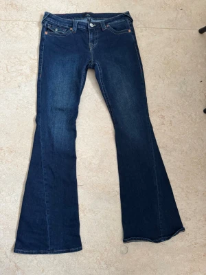 True Religion Joey Low Rise Flare Jeans - Mörkblå jeans från True Religion, modell Joey Low Rise Flare. Klassisk femficksdesign med kontrastsömmar och snygga detaljer på bakfickorna. Byxorna har låg midja och utsvängda ben för en retroinspirerad look. Perfekta för dig som gillar statement-jeans.