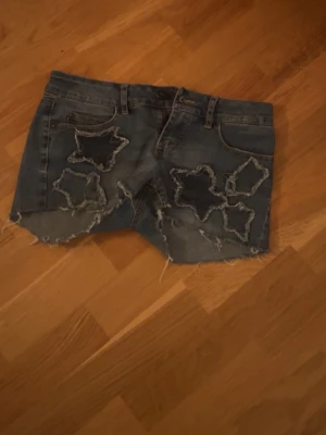 Blå jeansshorts med insydda stjärnor från ONLY - Säljer ett par blå jeansshorts från ONLY med råa kanter och slitna stjärnformade detaljer framtill. Säljer pga att de är för små.