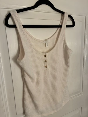 Vit ribbad linnetopp från H&M - Säljer en vit ribbad linnetopp från H&M i storlek M. Toppen har rund halsringning och tre snygga guldfärgade knappar framtill. Perfekt för varma dagar och enkel att matcha med jeans eller kjol.