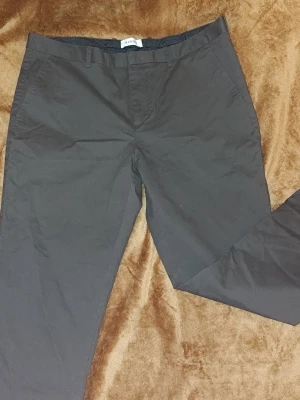 Mörkbruna chinos från Sandö - Snygga mörkbruna chinos från Sandö med klassisk rak passform. Byxorna har bälteshällor, snedställda fickor fram och två bakfickor med knapp. Tillverkade i bomull för en skön känsla och stilren look.