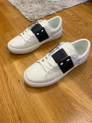 Valentino Garavani sneakers - Snygga vita sneakers från Valentino Garavani med bred svart läderdetalj över snörningen och coola nitar på hälen. Skorna har rund tå, platt sula och klassisk snörning. Perfekta för dig som vill ha en stilren men ändå unik look.