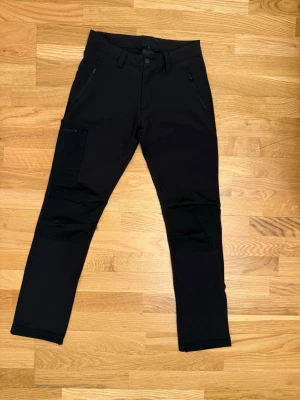 Svarta cargopants med dragkedjor - Säljer ett par svarta cargopants med snygga dragkedjor vid fickorna och förstärkta partier på knäna. Byxorna har en modern, tapered passform och är gjorda i ett slitstarkt syntetmaterial. Perfekta för dig som gillar funktionella och stilrena byxor.