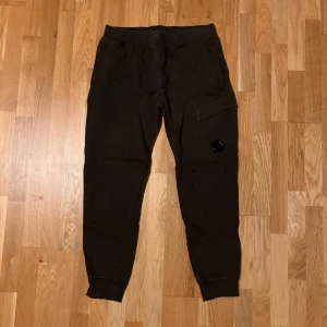 Brun/gröna mjukis cargopants från C.P. Company - Mörkbruna cargopants från C.P. Company med klassisk linsdetalj på benfickan. Byxorna har resår i midjan och vid bensluten, samt flera fickor för praktisk förvaring. Tillverkade i mjuk bomull för skön känsla och avslappnad stil.