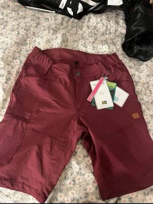 Vinröda shorts från Dobsom, strl 34 - Snygga vinröda shorts från Dobsom i storlek 34. De har praktiska fickor, bälteshällor och dragkedjor bak. Materialet är lätt och snabbtorkande, perfekt för aktiva dagar. Klassisk passform och stilren design.