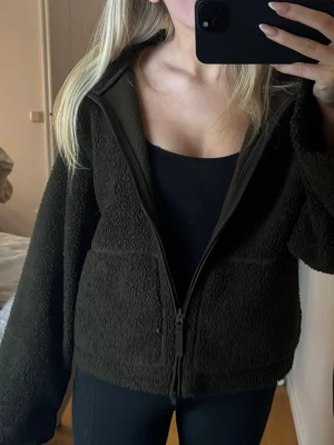 Mörkgrön teddyjacka med dragkedja - Mysig mörkgrön teddyjacka med hög krage och hel dragkedja framtill. Jackan har två stora fickor och en loose passform som ger en chill vibe. Perfekt för kalla dagar när du vill vara både varm och snygg.