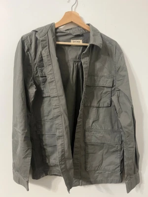 Grå overshirt från East West - Snygg grå overshirt från East West med fyra stora fickor framtill och klassisk krage. Jackan är tillverkad i bomull och har en avslappnad passform som passar perfekt till lager på lager. Enkel och stilren design som funkar till det mesta. Storlek S men passar även M