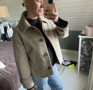 Beige kappa  - Säljer en beige kappa i ullblandning med klassisk krage och stora knappar framtill. Jackan har en rak passform och långa ärmar med knappdetalj vid mudden, vilket går att styla med klänning jeans eller kostymbyxor. Perfekt för dig som gillar stilren och enkel desig! Priset kan diskuteras 