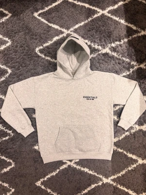 Fear of God Essentials hoodie - Svart hoodie med huva och känguruficka. Stilren design med diskret Essentials-logga på bröstet. Mjuk och bekväm passform, perfekt för vardagsbruk.Jag väger 70KG och är 180CM och den passar mig perfekt.