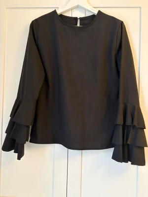 Svart blus med volangärmar - Snygg svart blus med rund halsringning och långa ärmar med flera lager volanger. Perfekt för dig som vill ha en stilren men ändå unik look. Blusen har en enkel siluett och är lätt att matcha med olika outfits.
