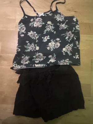 Blommig linnetopp med tunna axelband och snygga shorts  - Snygg svart linnetopp med blommigt mönster i lila och vitt. Toppen har tunna axelband och är perfekt för varma dagar. Lätt och luftig känsla, passar till både jeans och kjol. Säljer även shortsen de följer med 