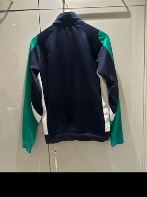 Lacoste Sport vindjacka blå/grön S - Snygg vindjacka från Lacoste Sport i mörkblått med gröna ärmar och vita detaljer längs sidorna. Jackan har dragkedja framtill, klassisk passform och hög krage. Perfekt för en sportig look och funkar året runt.