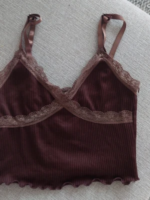 Brun ribbad linnetopp med spets H&M - Supersöt brun ribbad linnetopp från H&M Divided i storlek XS. Toppen har tunna satinband som axelband och fina spetsdetaljer längs kanterna. Perfekt för lager-på-lager eller som statement-piece till jeans eller kjol.