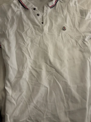 Vit Moncler pikétröja S - Klassisk vit pikétröja från Moncler i storlek S. Tröjan har krage med röd och marinblå rand, tre knappar framtill och Moncler-logga broderad på bröstet. Tillverkad i bomull för en skön och luftig känsla. Perfekt till jeans eller shorts.