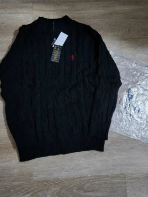 Svart stickad tröja Polo Ralph Lauren - Kabelstickad svart tröja från Polo Ralph Lauren med klassisk röd logga broderad på bröstet. Tröjan har rund halsringning, ribbade muddar och är långärmad. Perfekt för dig som gillar stilrena och tidlösa plagg.