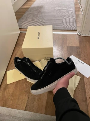 Svarta sneakers från Axel Arigato - Säljer ett par svarta sneakers från Axel Arigato med vit platt sula och dragkedja på ovansidan. Skorna är i mocka med detaljer i skinn och har en rund tå. Medföljer originalkartong och dustbag. Perfekt för dig som gillar stilrena och moderna sneakers.storlek 40 passar 41 hör av er för mer bilder obs box medföljer ej