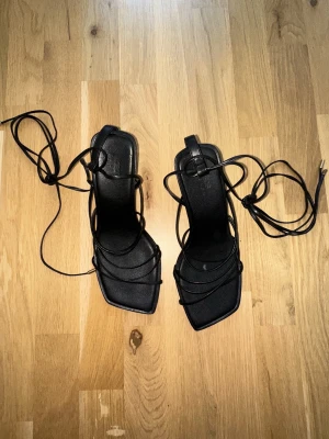 Svarta klackar med snörning från ASOS - Säljer ett par svarta klackskor från ASOS DESIGN med tunna remmar och snörning som går att vira runt benet. Skorna har fyrkantig tå och låg klack, tillverkade i syntetmaterial. Perfekta för dig som gillar minimalistisk och trendig stil. Använda en gång. Från ASOS wide-fit serie så passar bra om du har lite bredare fötter.