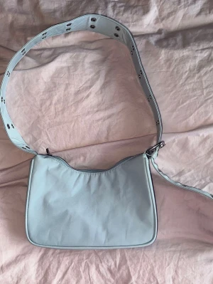 Ljusblå axelväska från weekday - Säljer en ljusblå axelväska med bred justerbar rem. Väskan har en enkel, stilren design och stängs med dragkedja. Materialet är slätt och väskan har ett innerfack med dragkedja för småsaker. Perfekt för dig som vill ha en cool och trendig accessoar. Bredd är 22 cm och höjd är 13 cm