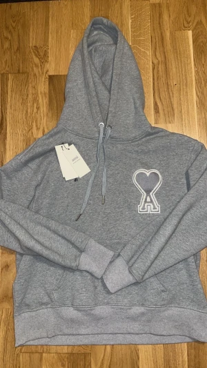 Grå hoodie från Ami Paris  - Säljer en grå hoodie från Ami Alexandre Mattiussi med stor broderad hjärtlogo på bröstet. Tröjan har huva med snörning, ribbade muddar och känguruficka. Materialet är mjuk bomull och passformen är normal. Perfekt för dig som gillar stilrena detaljer.