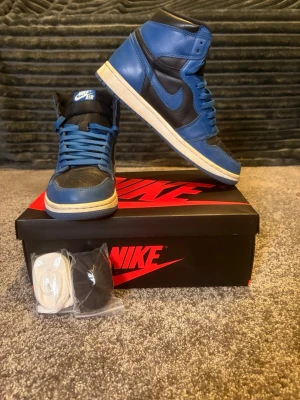 Nike Air Jordan 1 High blå/svarta - Säljer ett par Nike Air Jordan 1 High i blått och svart skinn. Klassisk siluett med rund tå, blå snörning och vit mellansula. Kommer med extra skosnören i vitt och svart. Perfekta för dig som vill sticka ut med ikoniska sneakers. Priset är ej spickat så hör av dig!