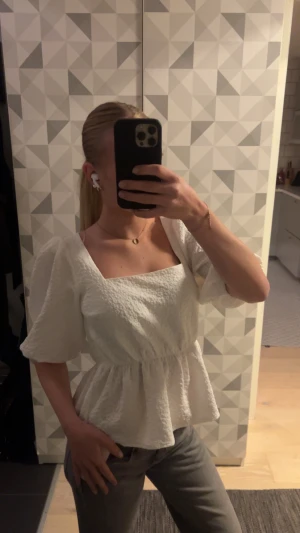 Vit blus med puffärm och struktur - Säljer en vit blus med fyrkantig ringning och puffiga korta ärmar. Toppen har en markerad midja och ett lätt strukturerat tyg som ger en cool look. Passformen är normal och blusen har en flowy volang nedtill. Perfekt för dig som gillar stilrena detaljer.