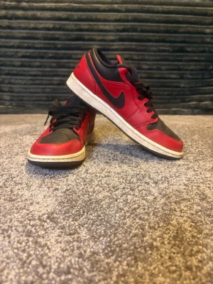 Nike Air Jordan 1 Low svart/röd - Säljer ett par Nike Air Jordan 1 Low i svart och rött skinn med klassisk svart swoosh på sidan. Skorna har svart snörning, vit sula och Air Jordan-logga broderad på hälen. Perfekta för dig som vill ha en ikonisk sneaker med street vibe. Priset är ej spickat 