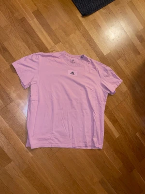 Rosa t-shirt från Adidas XL - Säljer en ljusrosa t-shirt från Adidas i storlek XL. T-shirten har klassisk passform, korta ärmar och rund halsringning. Adidas-loggan sitter tryckt på bröstet. Tillverkad i mjuk bomull som känns skön mot huden.