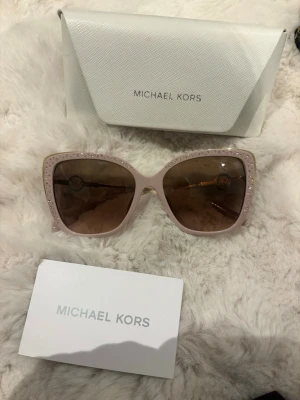 Rosa Michael Kors solglasögon - Säljer ett par snygga solglasögon från Michael Kors i ljusrosa med guldiga detaljer och glittriga sidor. Stora bågar med bruna glas och MK-logga på sidorna.
