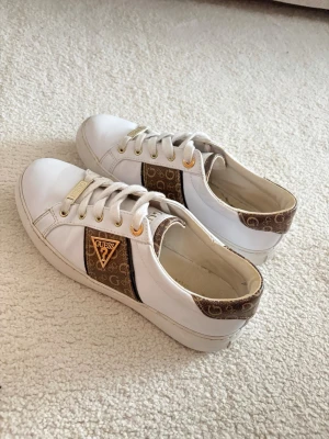 Vita sneakers från Guess med logga - Snygga vita sneakers från Guess med bruna detaljer och logomönster på sidor och häl. Skorna har guldfärgade metalldetaljer och klassisk snörning. Materialet är skinn och syntet, och de har en platt sula samt rund tå. Perfekta för dig som gillar stilrena men ändå unika sneakers.