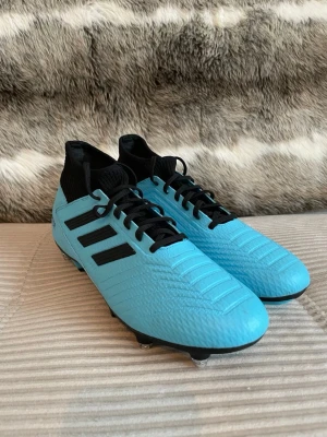 Adidas Predator blå fotbollsskor - Säljer ett par Adidas Predator fotbollsskor i en snygg ljusblå färg med svarta detaljer och snörning. Skorna har klassiska tre svarta ränder på sidan, högre strumpliknande skaft och dobbar i metall och plast. Perfekta för dig som vill sticka ut på planen.